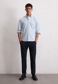 Camisa de botones a rayas en azul claro y blanco con un bolsillo en el pecho, combinada con pantalones y zapatos negros, sobre un fondo neutro.