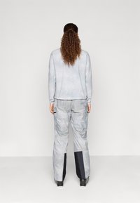 Cilvēks ar gariem, čirkainiem matiem, sasietiem ponijā, valkā gaiši pelēku jaku un bikses ar melniem pleķiem apakšējā kāju daļā, stāv pretēji.