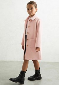 Abrigo de lana rosa con cuello y cinco botones de madera, con un bolsillo de parche. Llevado sobre una blusa blanca y combinado con botas negras de tobillo.