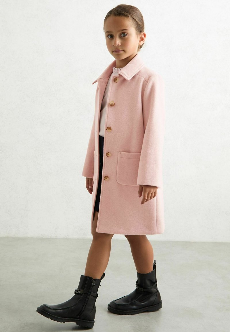 Abrigo de lana rosa con cuello y cinco botones de madera, con un bolsillo de parche. Llevado sobre una blusa blanca y combinado con botas negras de tobillo.