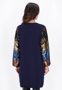 Lang marineblauwe jurk met lange mouwen van sequins in zwart, brons en een blauwe gradient. Zachte textuur en een ontspannen pasvorm. Geen zichtbare sluitingen.