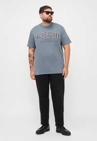 Grå bomull t-skjorte med "JACK & JONES" logo i svart, kombinert med svarte bukser og sorte sko, båret av en person med solbriller.