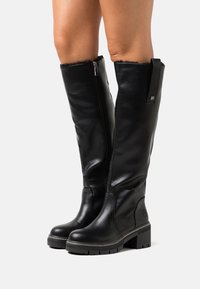Bottes noires montantes en cuir lisse, avec un extérieur texturé, une fermeture éclair sur le côté et une semelle robuste et épaisse avec un accent métallique subtil.