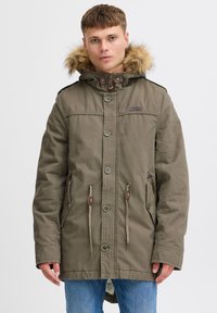Parka olive avec capuche en fourrure, fermetures à pression et éclair, cordons à la taille et plusieurs poches.