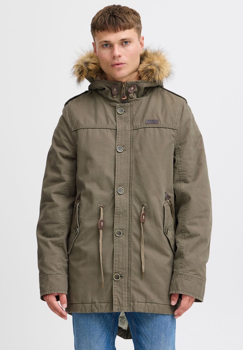 Parka olive avec capuche en fourrure, fermetures à pression et éclair, cordons à la taille et plusieurs poches.