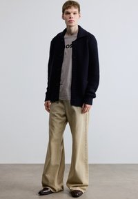 Tengerészkék kötött kardigán széles gallérral, gombos rögzítéssel és zsebekkel, szürke póló és bézs bőszárú nadrág fölött, sötét cipővel viselve.