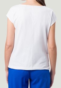 Top blanco de manga corta con escote redondeado, que presenta un detalle de costura en el centro de la parte posterior, combinado con pantalones azules.