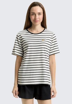 Kobieta o długich brązowych włosach, ubraną w białą koszulkę z czarnymi poziomymi paskami i małym żółtym detalem owocowym, w zestawieniu z czarnymi szortami.