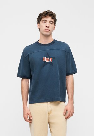 Joven con cabello rizado lleva una camiseta azul marino de gran tamaño con una estrella oscura y texto "USA", combinada con pantalones beige.