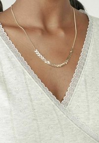 Goldene Halskette mit Chevron-Design, getragen über einem hellgrauen Oberteil mit Spitzenrand und einem subtilen Herzmuster.