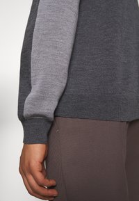 Grau und hellgrauer Strickpullover mit gerippten Bündchen, Rundhalsausschnitt und einer leicht lockeren Passform. Finger sind sichtbar und ruhen auf braunen Stoffhosen.