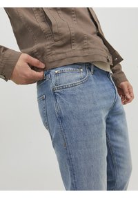 Person som bär ljusblå denimjeans och en brun jacka, justerar jackans fåll med vänster hand.