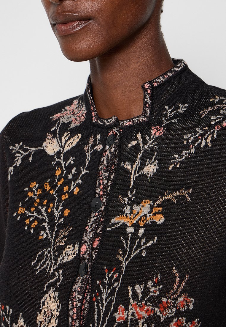Close-up van een persoon die een zwarte, knoopsluiting vest draagt met een meerkleurig gebreid bloemenpatroon en een ronde uitsnijding op de kraag.