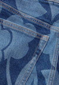 Vaaleansininen denimmateriaali, jossa tummansininen kukkakuviointi, näkyvine sauman yksityiskohtineen ja teksturoidun pinnan kanssa.