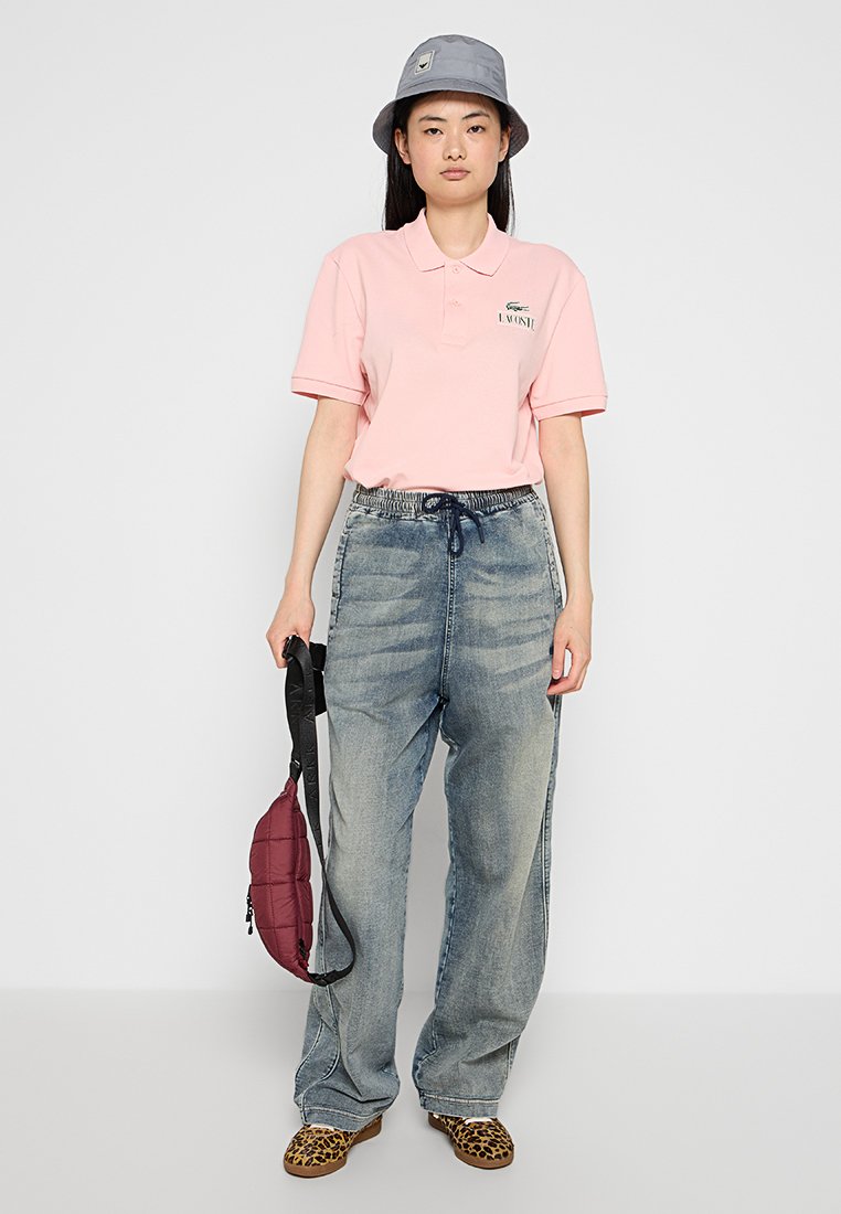 Kobieta w szarym kapeluszu typu bucket, różowej koszulce polo Lacoste, luźnych, wyblakłych dżinsach oraz butach w panterkę, trzymająca przy sobie burgundową, pikowaną torebkę.