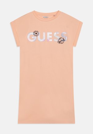 Robe t-shirt en coton pêche avec des manches courtes retroussées. Présente "GUESS" en lettres réfléchissantes et un détail de logo circulaire sur le devant.