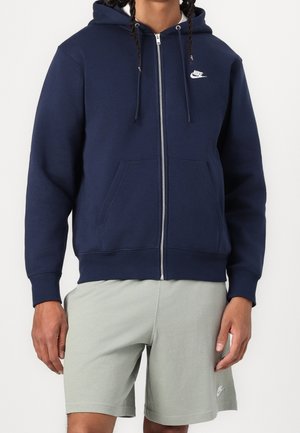 Personne portant un sweat à capuche zippé bleu marine Nike avec cordons et un short gris clair Nike, debout devant un fond uni.