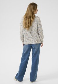 Vrouw met lang haar, gekleed in een multicolore gebreide trui en losse blauwe jeans, staande en met de rug naar de camera op een effen achtergrond.