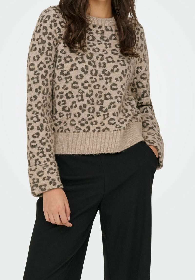 Personne portant un pull beige à manches longues imprimé léopard et un pantalon noir, une main dans la poche et l'autre détendue le long du corps.