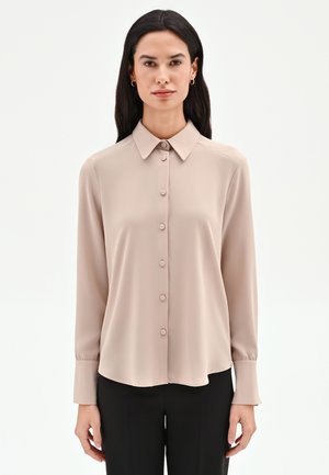 Donna che indossa una camicetta beige a maniche lunghe con colletto e bottoni, abbinata a pantaloni neri, in piedi davanti a uno sfondo bianco semplice.