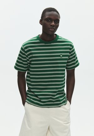 Ung mand med mørk hud iført en grøn og hvidstribet T-shirt og hvide shorts, stående med hænderne i lommerne mod en ensfarvet baggrund.