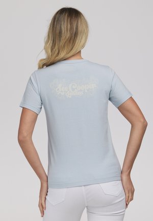 Kobieta o długich blond włosach ma na sobie jasno-niebieską koszulkę z krótkim rękawem z białym kwiatowym logo "Lee Cooper London" na plecach, w parze z białymi spodniami.