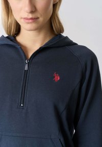 Felpa blu navy con colletto con zip e un piccolo logo rosso. Il materiale è morbido con una texture liscia e un design casual.