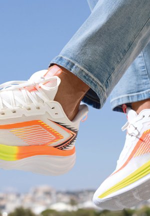 White athletic shoes with orange, yellow, and black accents worn with light blue jeans against a clear blue sky background.