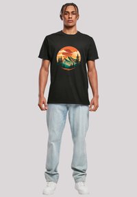 Black cotton t-shirt featuring a circular graphic of mountains and trees in orange, turquoise, and green hues. Model wears light blue jeans.