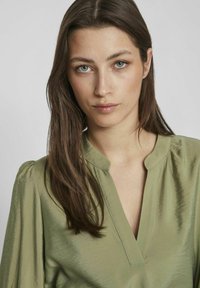 Olivgrön blus med V-ringning och plisserade puffärmar, tillverkad av slätt, glänsande tyg, med subtila sömnadsdetaljer.
