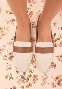 Mocassins en toile beige avec des accents en cuir marron, présentant un design plat et un bout pointu, placés sur une surface à motif floral.