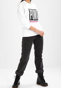 Felpa bianca con la scritta nera "PUMA" e accento rosa, abbinata a jeans neri arrotolati e stivali neri con lacci. Tessuto liscio, vestibilità casual.