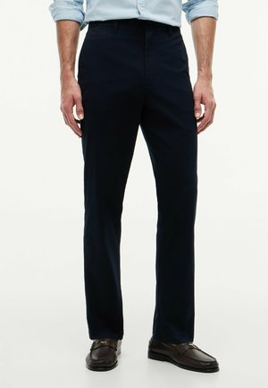 Mężczyzna w ciemnogranatowych spodniach, jasnoniebieskiej podwiniętej koszuli i ciemnobrązowych skórzanych mokasynach, stojący na białym tle.