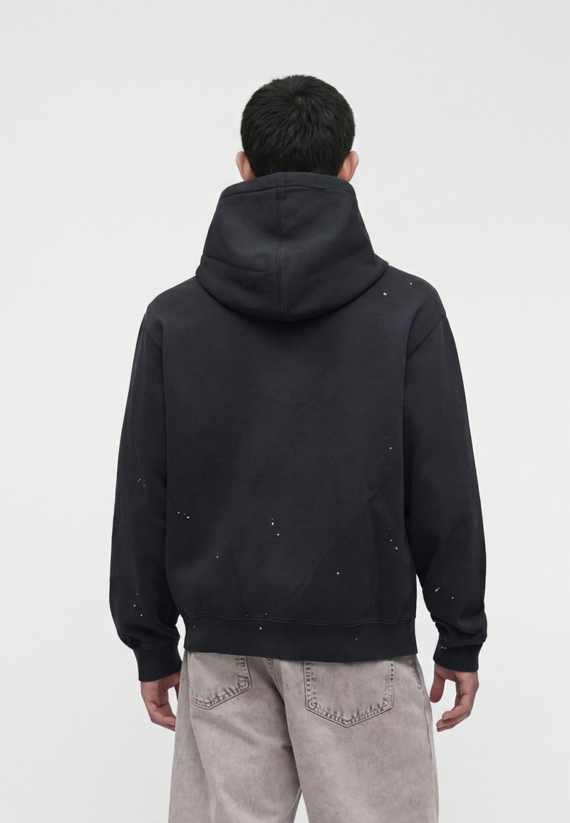 Homme de dos portant un sweat à capuche noir avec de petites éclaboussures blanches de peinture et un jean gris clair sur un fond blanc uni.