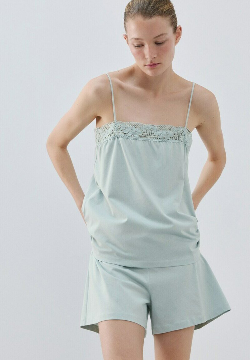 Femme portant un haut sans manches bleu clair avec bordure en dentelle et un short assorti, debout devant un fond clair uni.