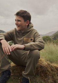 Hoodie w kolorze oliwkowym z mieszanki bawełny, z kieszenią z przodu, luźny krój i ściągacze na mankietach. W połączeniu z dopasowanymi spodniami dresowymi i czarnymi butami turystycznymi.