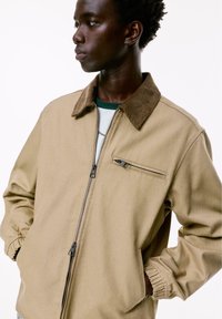 Giovane uomo in giubbotto beige con zip e colletto marrone, mani nelle tasche, che guarda di lato su uno sfondo bianco.