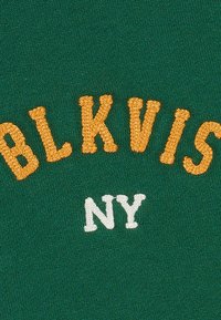 Zelená látka so vyšitým textom: „BLKVIS“ v oranžovej a „NY“ v bielej farbe. Písmená sú tučné a majú textúrovaný steh.