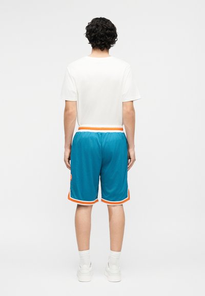 Hátulról látható férfi fehér pólóban, kék hálós kosárlabdazsákkal narancssárga szegéllyel, fehér bokazokniban és fehér tornacipőben.