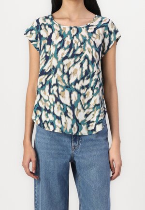 Femme portant un haut à manches courtes avec imprimé léopard abstrait en turquoise, blanc, beige et bleu marine, associé à un jean en denim bleu, sur un fond uni.