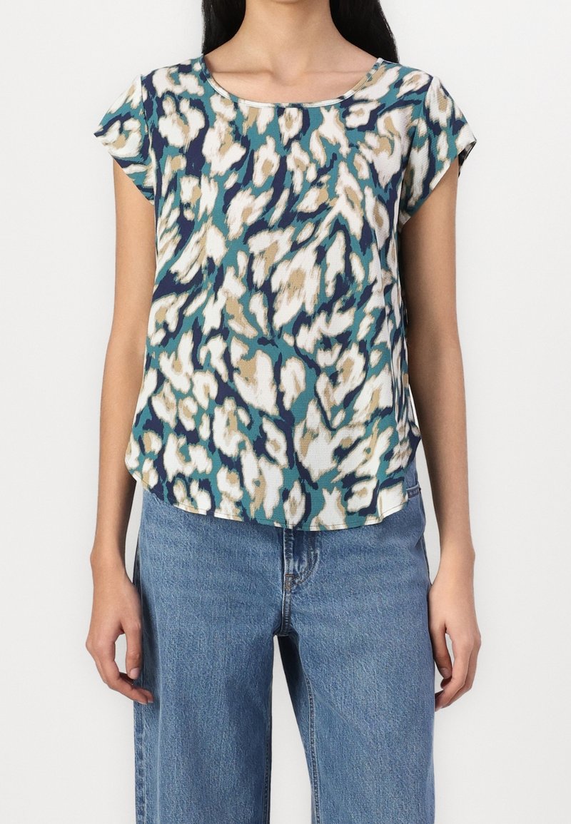 Femme portant un haut à manches courtes avec imprimé léopard abstrait en turquoise, blanc, beige et bleu marine, associé à un jean en denim bleu, sur un fond uni.