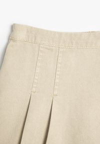 Beige geplooide stof met een gladde textuur. Zichtbare stiksels langs de tailleband en plooilijnen, die een gestructureerd ontwerp laten zien.