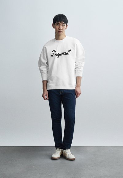 Λευκό φούτερ με μαύρο λογότυπο "Dsquared2", συνδυασμένο με σκούρο τζιν και μπεζ αθλητικά παπούτσια. Το μοντέλο στέκεται μπροστά σε ανοιχτό γκρι φόντο.