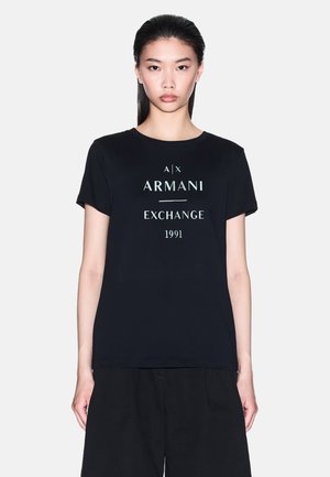 Donna con capelli lisci e scuri indossa una t-shirt nera Armani Exchange con scritta bianca e pantaloni neri, in piedi davanti a uno sfondo chiaro e semplice.