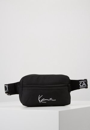 Sac banane noir avec une poche zippée à l’avant, une sangle de logo ajustable et des lettres blanches signature, exposé sur un piédestal blanc.