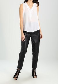 Witte mouwloze blouse met een V-halslijn, gecombineerd met zwarte leren broeken en zwarte veterschoenen tot de enkel, staande op een lichte achtergrond.