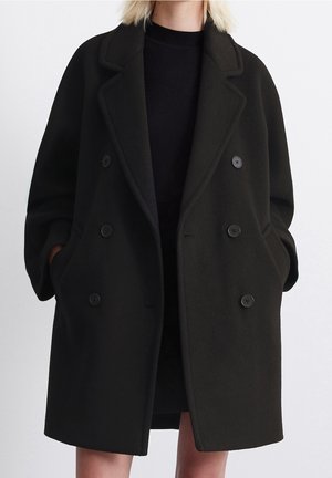 Kvinde iført en sort oversized dobbeltradet uldfrakke med notchkraver og hænderne i lommerne, foran en ensfarvet baggrund.