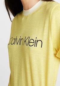Žluté pruhované tričko s klasickým kulatým výstřihem a krátkými rukávy. Na hrudi má výrazné černé logo "Calvin Klein".