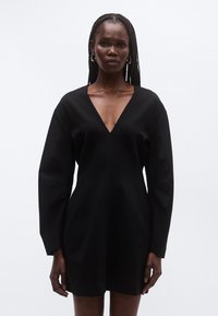 Μαύρο μίνι φόρεμα με βαθύ V ντεκολτέ, δομημένα μακριά μανίκια και εφαρμοστή σιλουέτα, φτιαγμένο από λείο, υφασμάτινο ύφασμα με υφή.