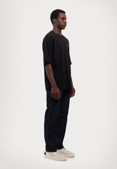 T-shirt noir oversized à manches courtes, associé à un jean en denim bleu foncé et des baskets beiges avec des accents noirs.